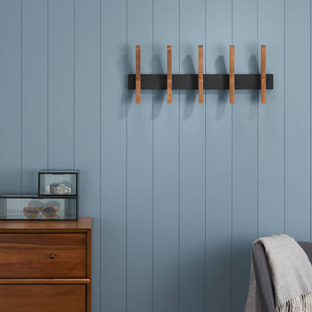 A modern wall-mounted coat rack with wooden pegs is centered on a blue paneled wall. A dresser, mirror, clock, and chair are visible in the surrounding room.