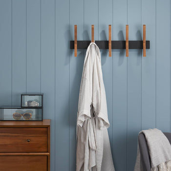 A horizontal black rack with five retractable wooden pegs is mounted on a light blue paneled wall. A white bathrobe hangs from the second peg from the left.