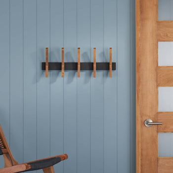 A Mid-Century Coat Rack mounted on a blue wall. The coat rack features a black base with wooden pegs, positioned next to a wooden door with frosted glass panels.