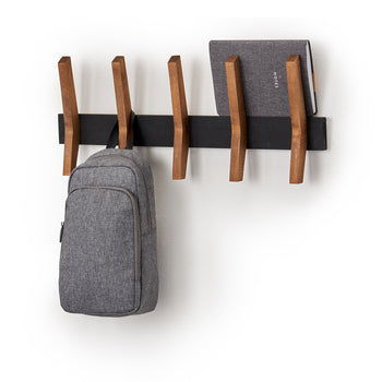 A minimalist black and wooden coat rack is mounted on a white wall, holding a gray backpack and a closed gray notebook. The coat rack features five evenly spaced, angled wooden pegs extending from a sleek black horizontal bar.