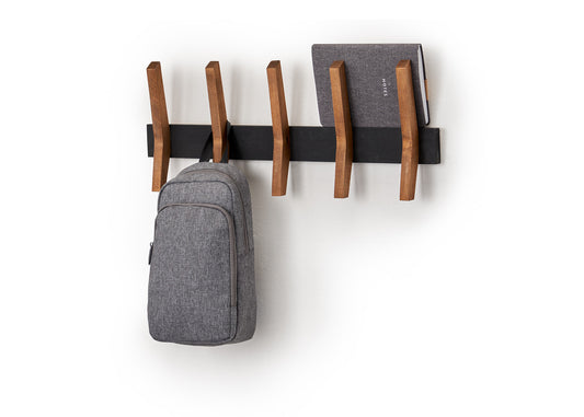 A minimalist black and wooden coat rack is mounted on a white wall, holding a gray backpack and a closed gray notebook. The coat rack features five evenly spaced, angled wooden pegs extending from a sleek black horizontal bar.
