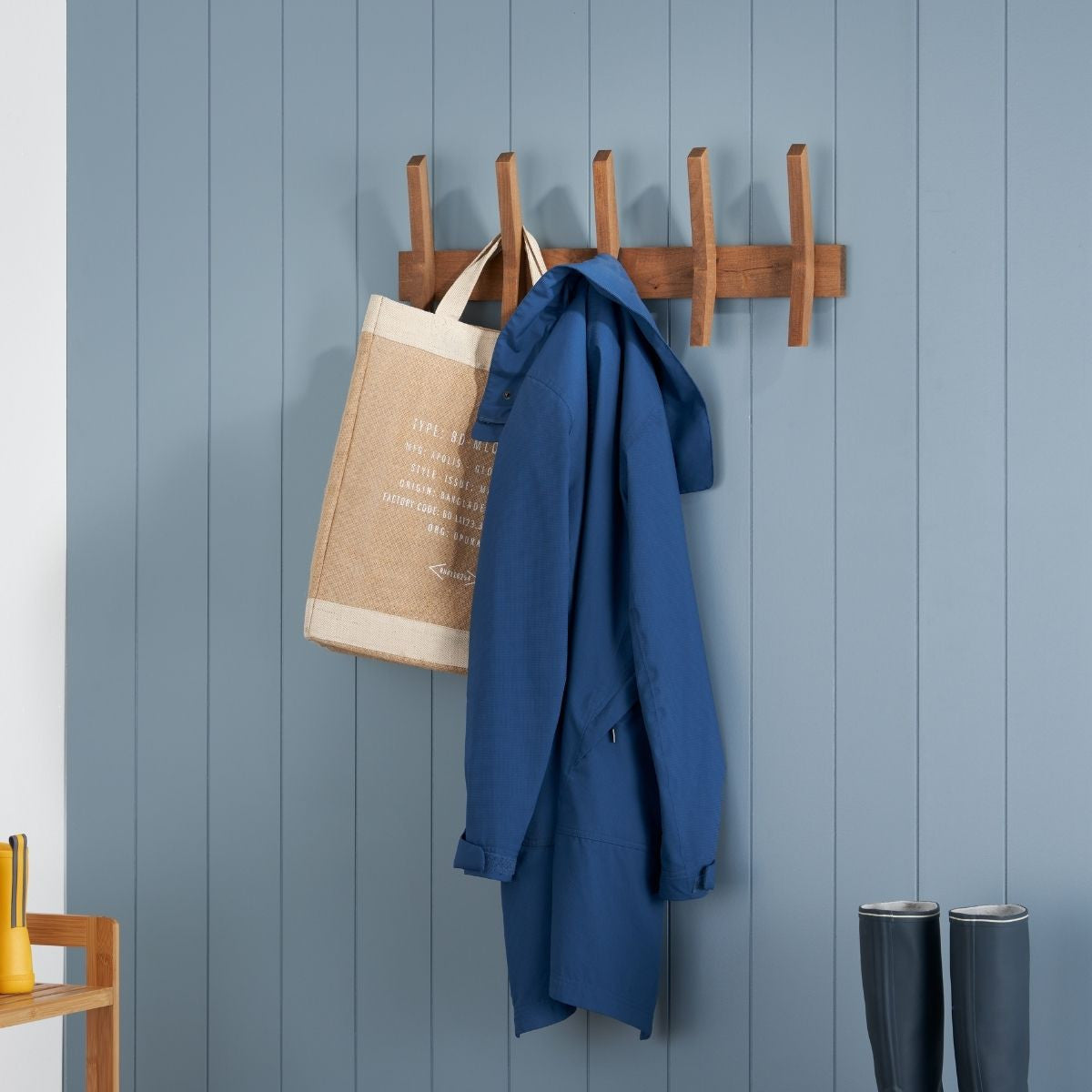 A wooden five-hook rack is mounted on a blue paneled wall, holding a beige tote bag on the leftmost hook and a folded blue jacket on the center hook. Below the rack, a pair of dark gray rubber boots stand against the wall.