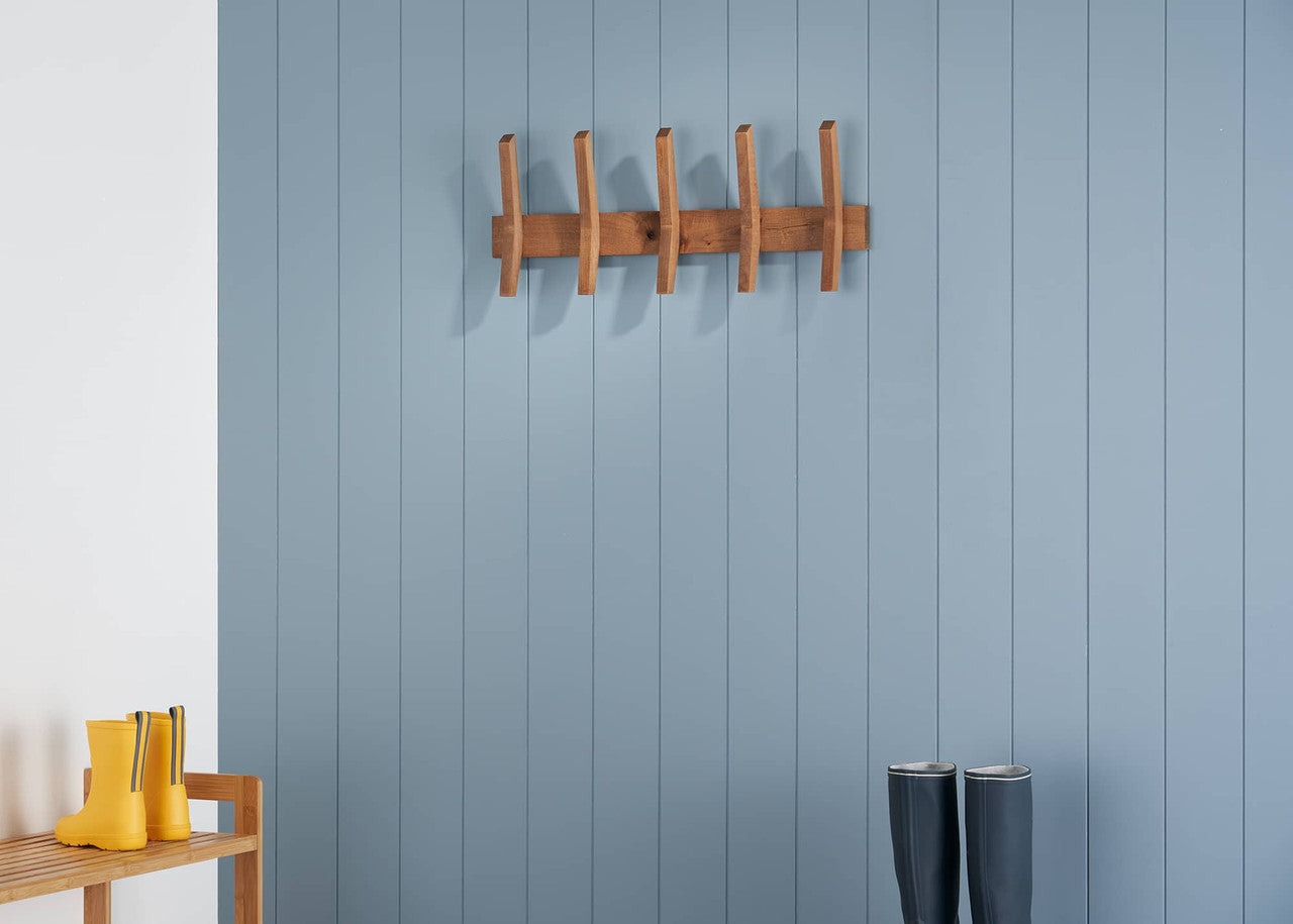 A wooden coat rack with five vertical pegs and two horizontal supports is mounted on a blue, vertically paneled wall. Below, a wooden shoe rack holds bright yellow rain boots on the left, and a pair of dark gray rain boots stand on the floor to the right.