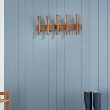 A wooden coat rack with five vertical pegs and two horizontal supports is mounted on a blue, vertically paneled wall. Below, a wooden shoe rack holds bright yellow rain boots on the left, and a pair of dark gray rain boots stand on the floor to the right.