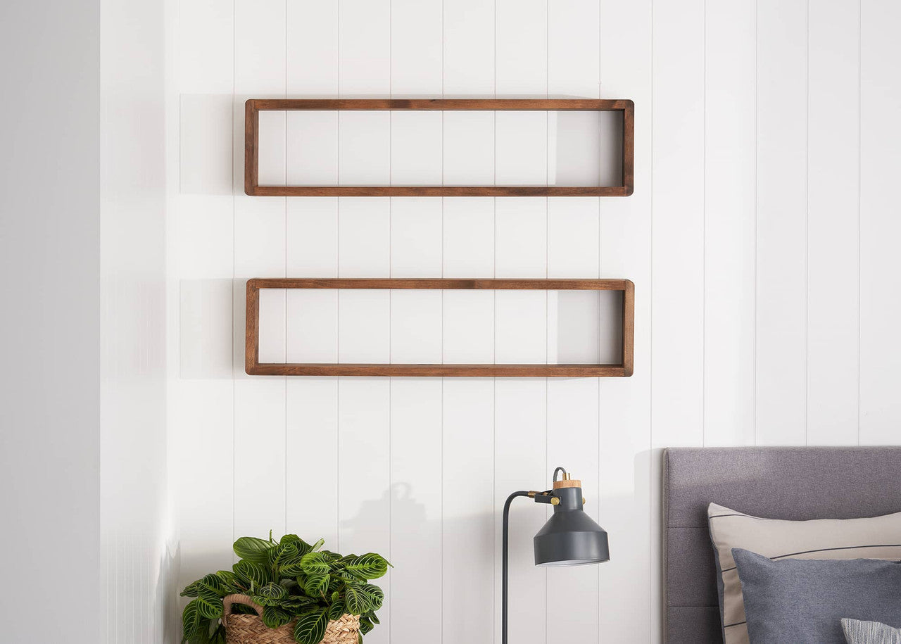 Two rectangular, open-backed wooden shelves are mounted on a white paneled wall. Below, a leafy green plant sits in a woven basket near a black desk lamp and the corner of a gray upholstered headboard.