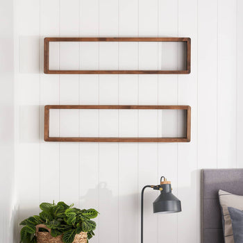 Two rectangular, open-backed wooden shelves are mounted on a white paneled wall. Below, a leafy green plant sits in a woven basket near a black desk lamp and the corner of a gray upholstered headboard.