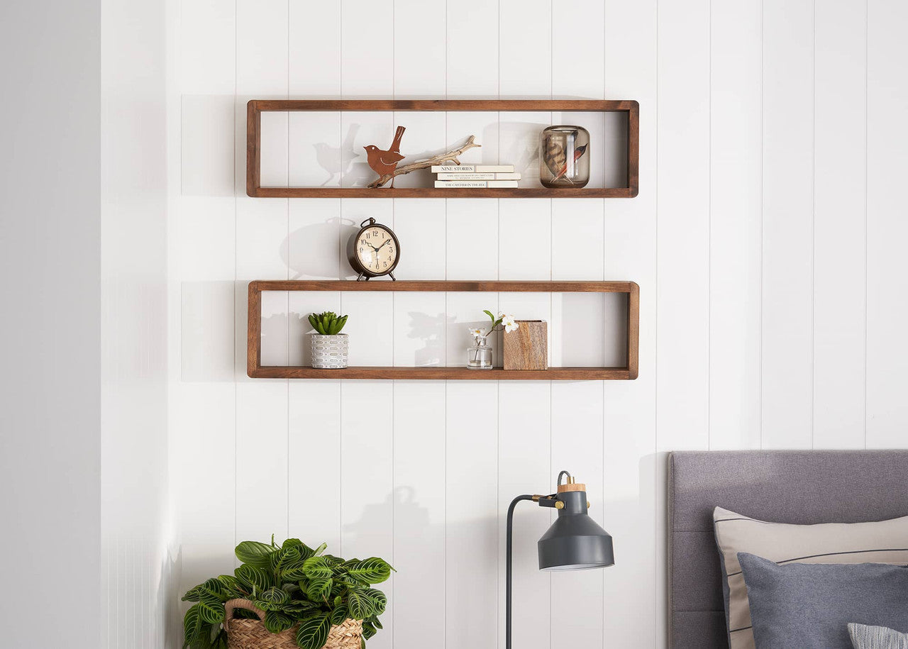 Two long, rectangular wooden floating shelves are mounted on a white paneled wall. The top shelf holds a decorative bird, a stack of small books, and a glass jar, while the lower shelf displays a small potted plant, a vase with greenery, and a wooden container.