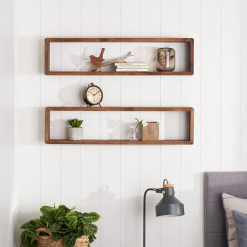 Two long, rectangular wooden floating shelves are mounted on a white paneled wall. The top shelf holds a decorative bird, a stack of small books, and a glass jar, while the lower shelf displays a small potted plant, a vase with greenery, and a wooden container.