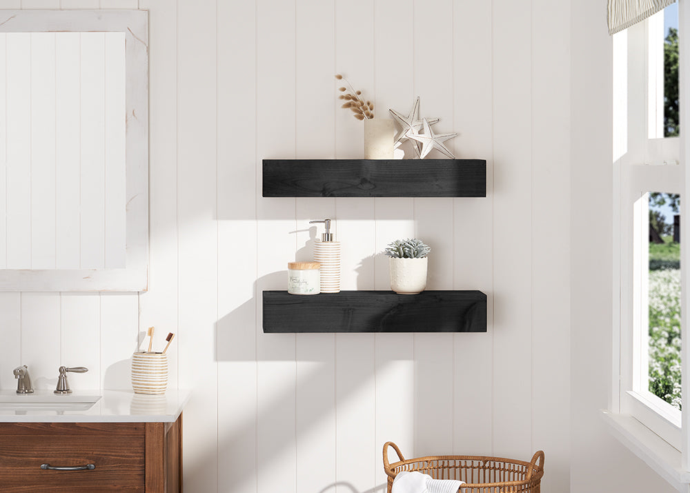 Two floating black wooden shelves are mounted on a white shiplap bathroom wall, decorated with coastal-themed items like starfish and a small succulent. The scene is brightened by natural sunlight from a nearby window, positioned above a wooden vanity with a white countertop.
