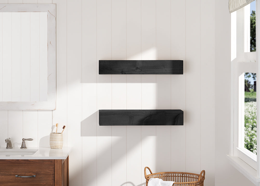 Two floating black wooden shelves are mounted on a white shiplap wall in a brightly lit bathroom. The scene includes a partial view of a wooden vanity with a white countertop on the left and a woven basket on the floor.