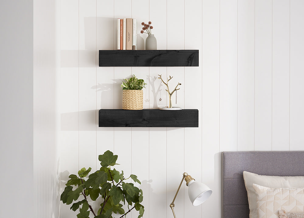 Two black floating shelves are mounted on a white, vertically paneled wall, displaying various decorative items. The top shelf holds books and a small vase with dried flowers, while the bottom shelf features a potted plant, a jewelry stand, and a small tray.