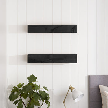 Two dark wood floating shelves are mounted vertically on a white shiplap wall above a bedside area with a lamp and a green plant. The scene features a modern, minimalist aesthetic with clean lines and soft, natural lighting.