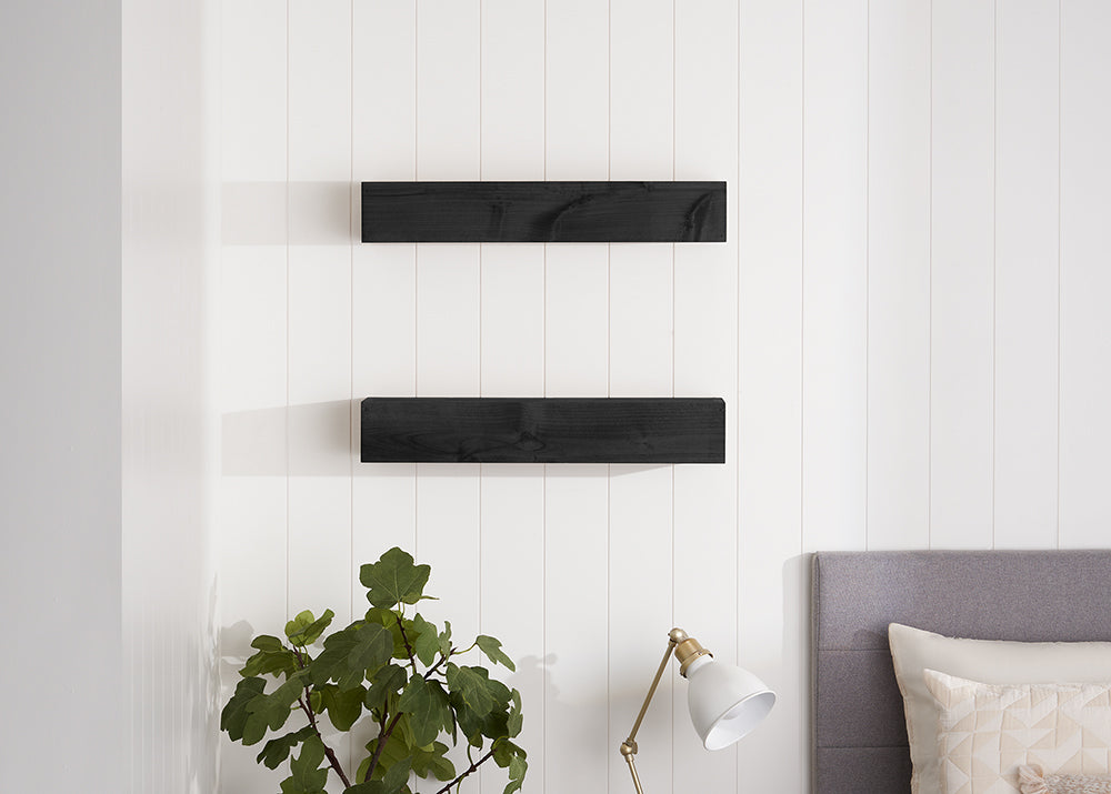 Two dark wood floating shelves are mounted vertically on a white shiplap wall above a bedside area with a lamp and a green plant. The scene features a modern, minimalist aesthetic with clean lines and soft, natural lighting.