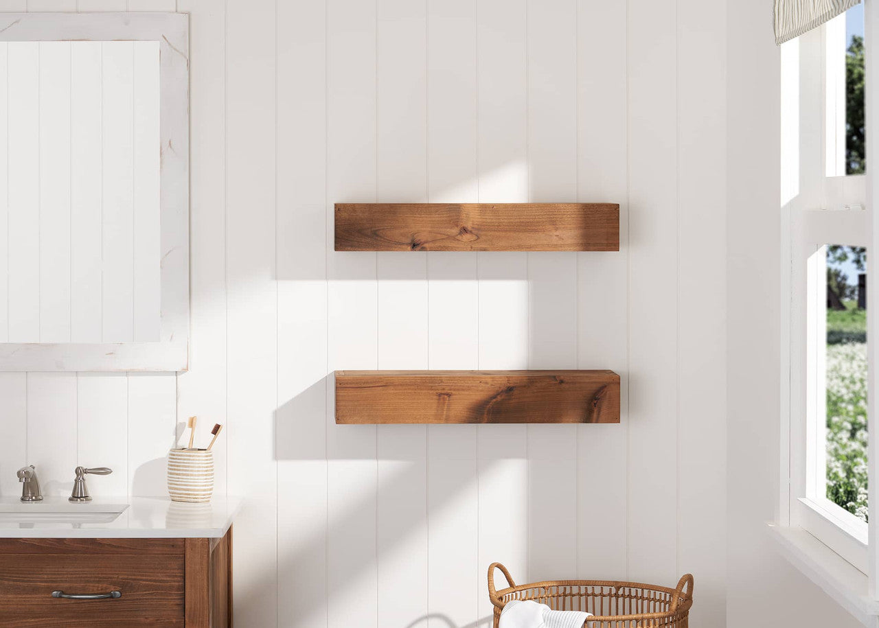 A bathroom with a white shiplap wall features two floating wooden shelves mounted above a wicker basket. A vanity with a white countertop, a mirror with a distressed wood frame, and a striped ceramic toothbrush holder is visible on the left.