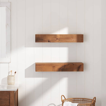 A bathroom with a white shiplap wall features two floating wooden shelves mounted above a wicker basket. A vanity with a white countertop, a mirror with a distressed wood frame, and a striped ceramic toothbrush holder is visible on the left.