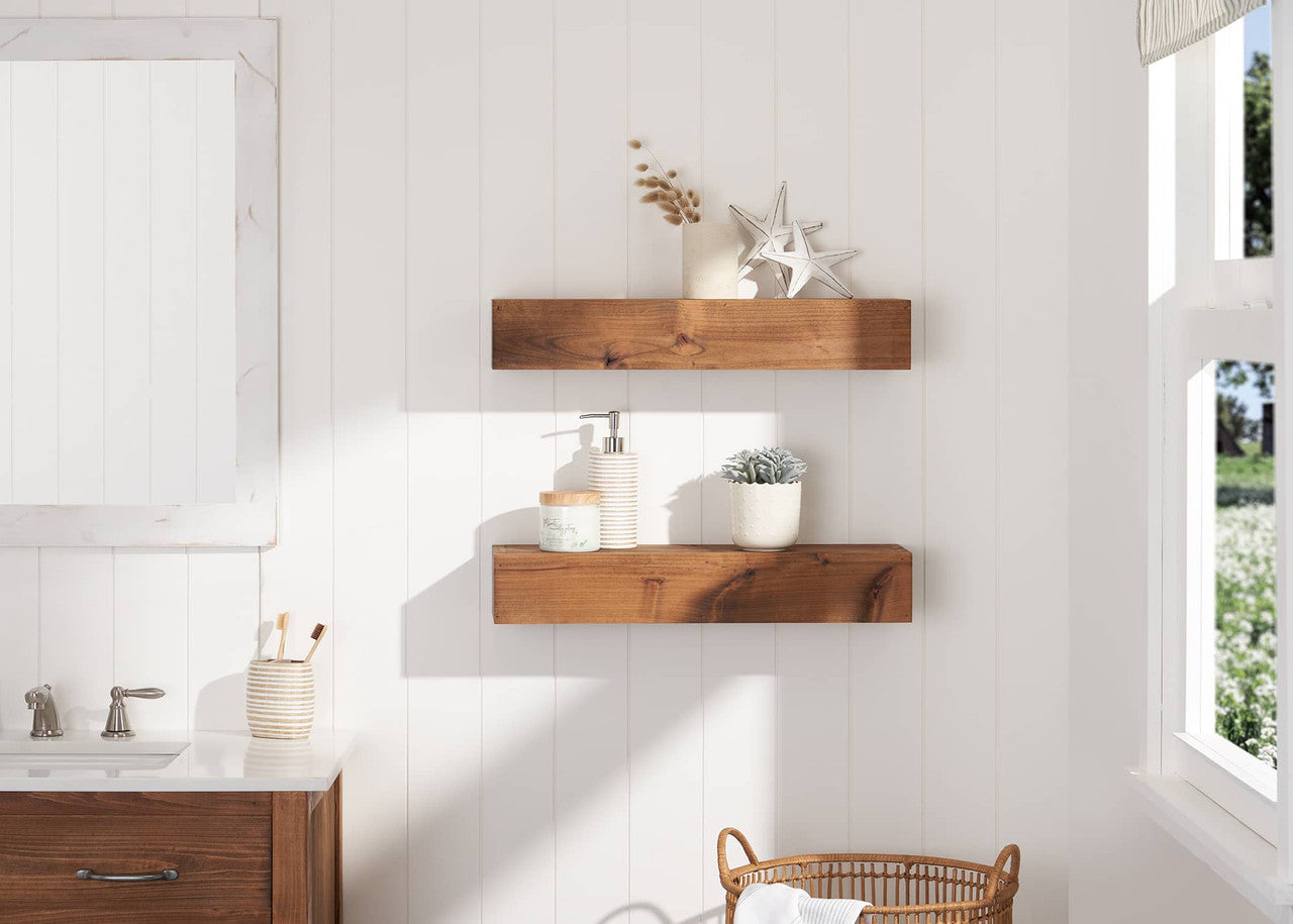 A bathroom with white paneled walls features two wooden floating shelves holding decorative items like a potted plant, soap dispenser, and silver star ornaments. Natural light streams in from an open window, illuminating the space with a cozy, rustic aesthetic.