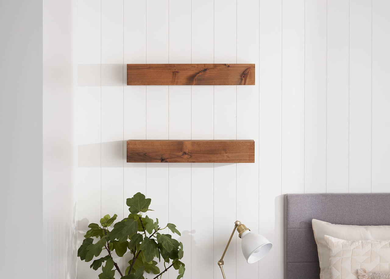 Two rectangular, light-brown wooden shelves are mounted flush against a white, vertically paneled wall.  A leafy green plant sits to the left on the floor, and a portion of a gray bed and headboard are visible on the right.