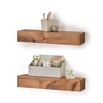 Two wooden floating shelves are mounted on a white wall, each holding office supplies and decorative items. The top shelf has a small container with pencils and a rubber band ball, while the bottom shelf holds an organizer with scissors, stationery, and a small gold jack decoration.