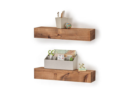 Two wooden floating shelves are mounted on a white wall, each holding office supplies and decorative items. The top shelf has a small container with pencils and a rubber band ball, while the bottom shelf holds an organizer with scissors, stationery, and a small gold jack decoration.