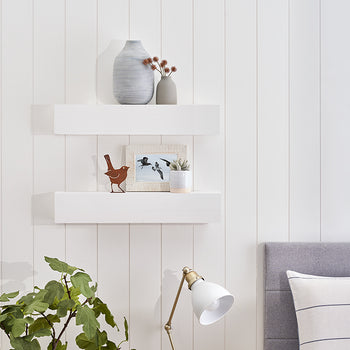 Two white floating shelves are mounted on a white vertical shiplap wall, decorated with vases, framed artwork, and a small wooden bird figurine. Below the shelves, a brass and white desk lamp illuminates a large green plant next to a grey upholstered headboard.