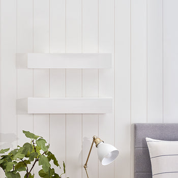 Two white floating shelves are mounted on a white vertical shiplap wall above a gray upholstered headboard. A green potted plant and a white desk lamp with brass accents sit on a surface in the foreground.