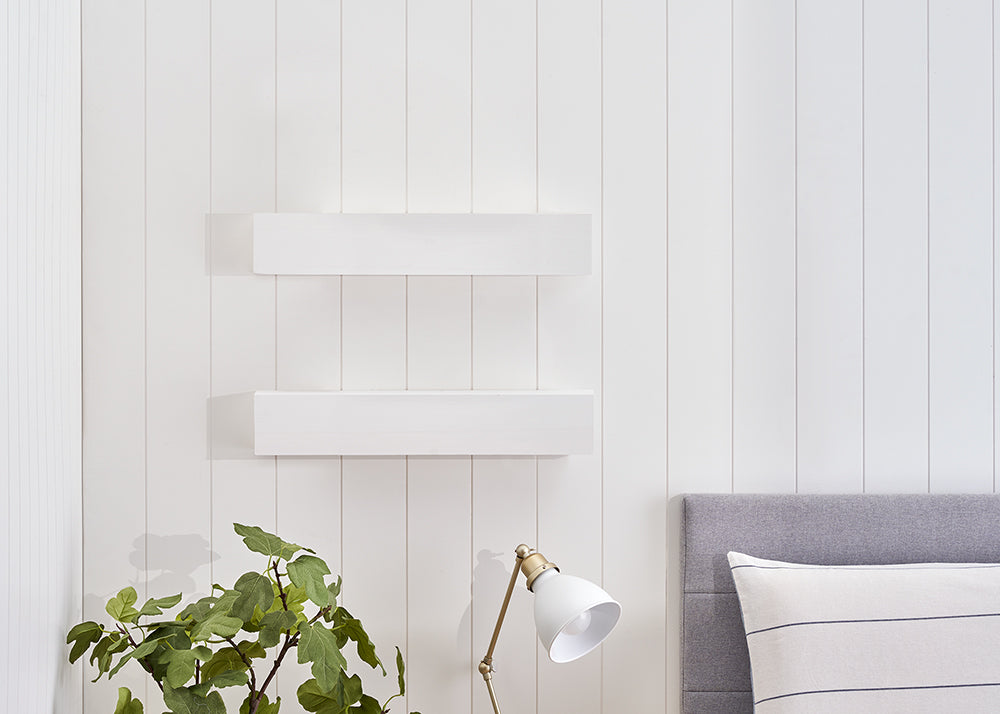 Two white floating shelves are mounted on a white vertical shiplap wall above a gray upholstered headboard. A green potted plant and a white desk lamp with brass accents sit on a surface in the foreground.