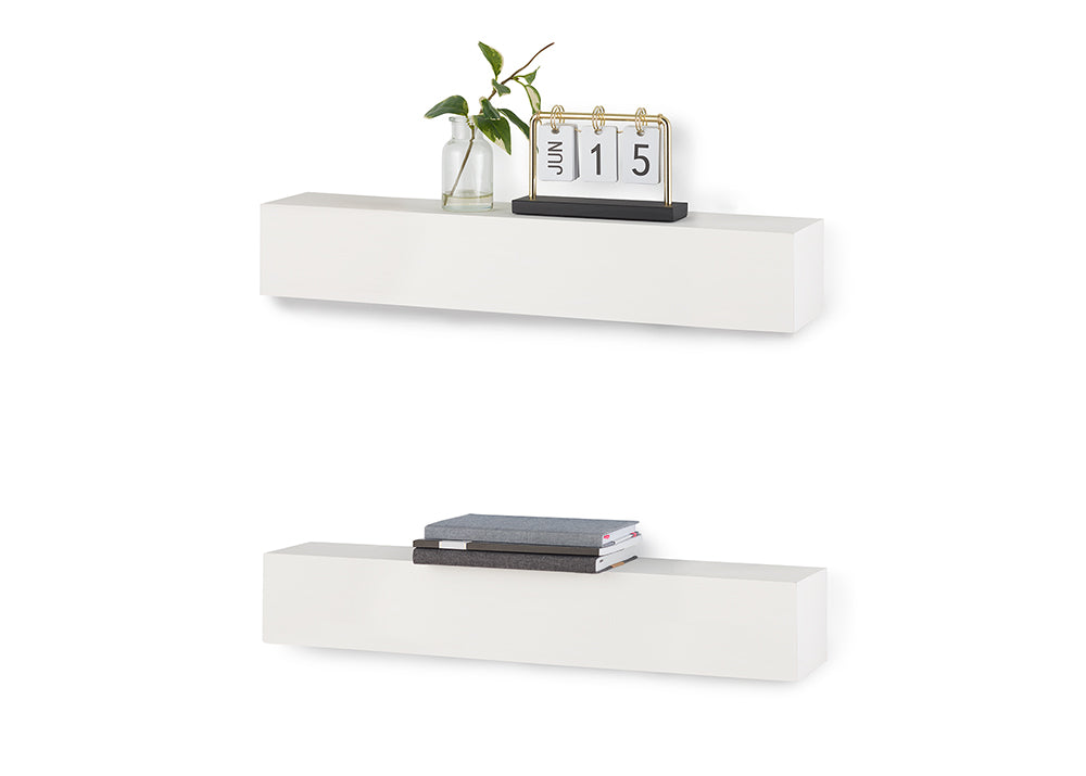 Two rectangular white floating shelves are mounted one above the other against a white background. The top shelf holds a small glass vase with a green sprig and a gold and black flip calendar displaying "JUN 15", while the bottom shelf holds a small stack of books.