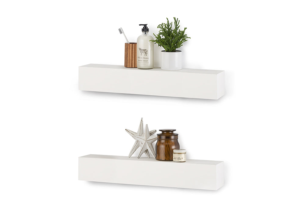 Two white floating shelves are mounted vertically on a white wall, styled with various bathroom and decorative items. The top shelf holds a wooden toothbrush holder, a bottle of hand soap, and a small potted plant, while the bottom shelf displays two decorative stars and two glass jars.