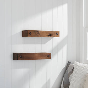 A pair of rustic wooden floating shelves with visible knots and metal hardware are mounted on a white paneled wall near a window. The cozy corner features neutral-toned pillows, and the window offers a view of a green field with white flowers.