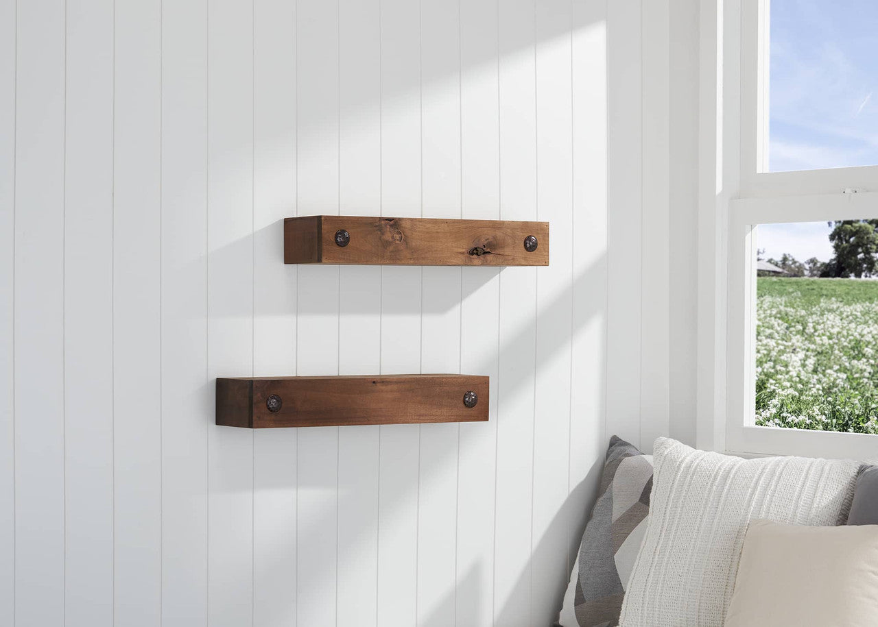 A pair of rustic wooden floating shelves with visible knots and metal hardware are mounted on a white paneled wall near a window. The cozy corner features neutral-toned pillows, and the window offers a view of a green field with white flowers.