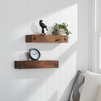 A cozy corner with two rustic wooden floating shelves mounted on a white paneled wall, decorated with a black bird figurine, a small potted plant with purple flowers, and a round black alarm clock. Sunlight streams in through an open window, revealing a lush green field with white flowers outside.