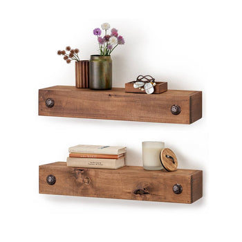 Two rustic wooden floating shelves are mounted on a white wall, each featuring two decorative metal studs on the front. The top shelf holds small vases with flowers, a wooden box with jewelry, while the bottom shelf has stacked books, a candle, and a small wooden container with a lid.