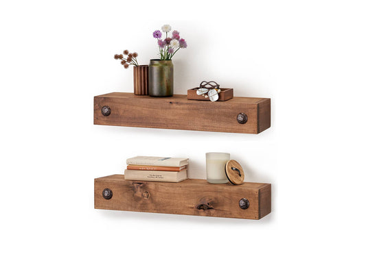 Two rustic wooden floating shelves are mounted on a white wall, each featuring two decorative metal studs on the front. The top shelf holds small vases with flowers, a wooden box with jewelry, while the bottom shelf has stacked books, a candle, and a small wooden container with a lid.