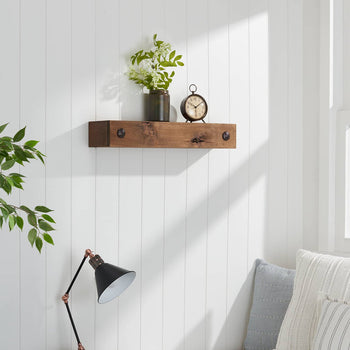 A medium-toned wood floating shelf with black metal brackets is mounted on a white shiplap wall in a well-lit room. The shelf displays a vintage-style clock and a dark vase with white blossoms, with a green leafy plant to the left and a gray and white striped pillow visible below.