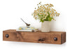 A wooden floating shelf holds a small stack of books titled "THE ART OF CIVILIZED CONVERSATION," with a small bird figurine perched on top, and a vase of white and yellow flowers. The shelf is rustic, made of thick wood with a natural edge and metal accents on the wall mounts.