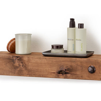 A farmhouse floating shelf is mounted on a white wall, holding a small tray with lotion and perfume bottles labeled 