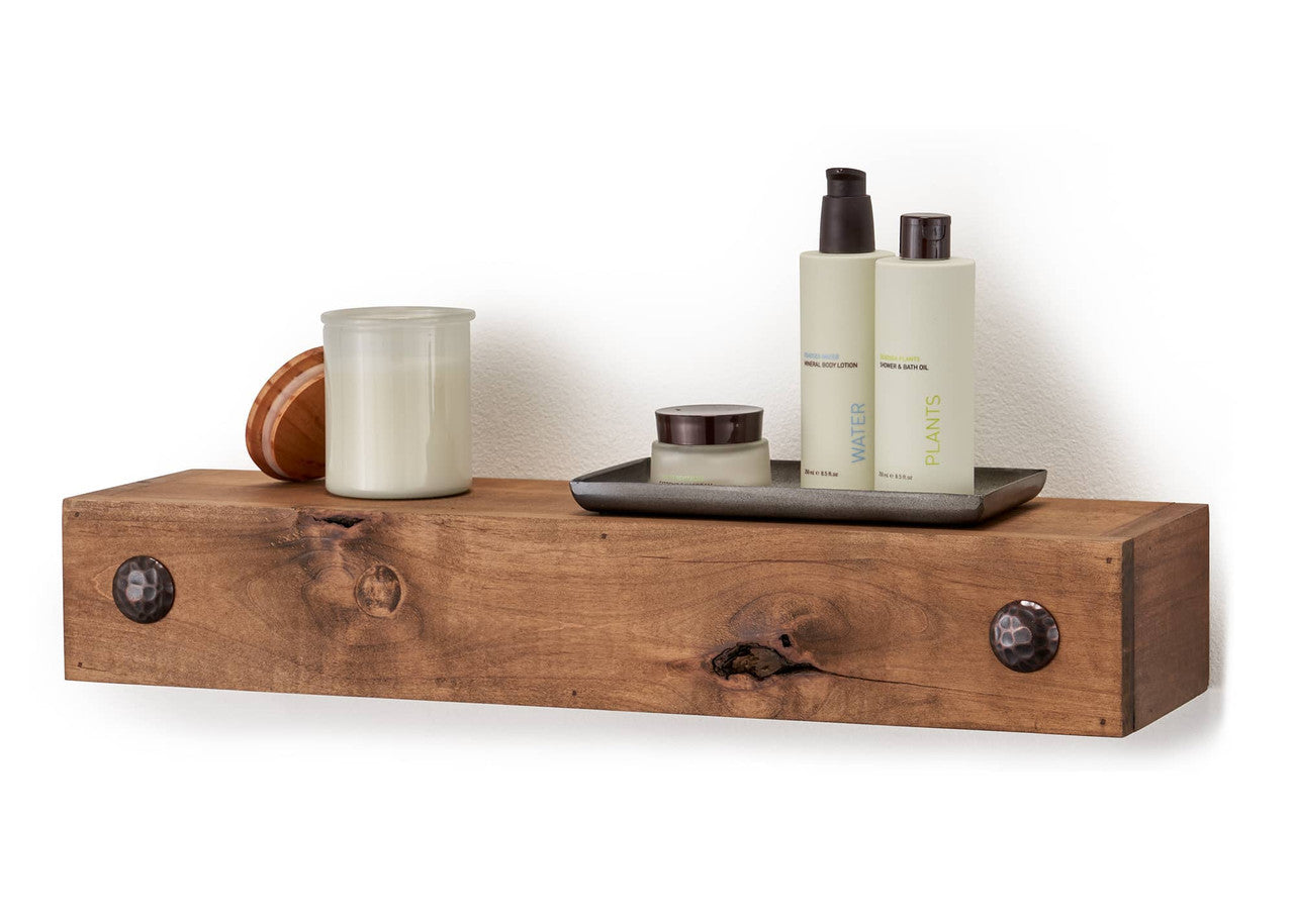 A farmhouse floating shelf is mounted on a white wall, holding a small tray with lotion and perfume bottles labeled "WATER" and "PLANTS," a candle in a glass jar with a wooden lid, and a small, round wooden object. The rustic shelf is made of wood with a natural finish and features two black metal brackets on either side.