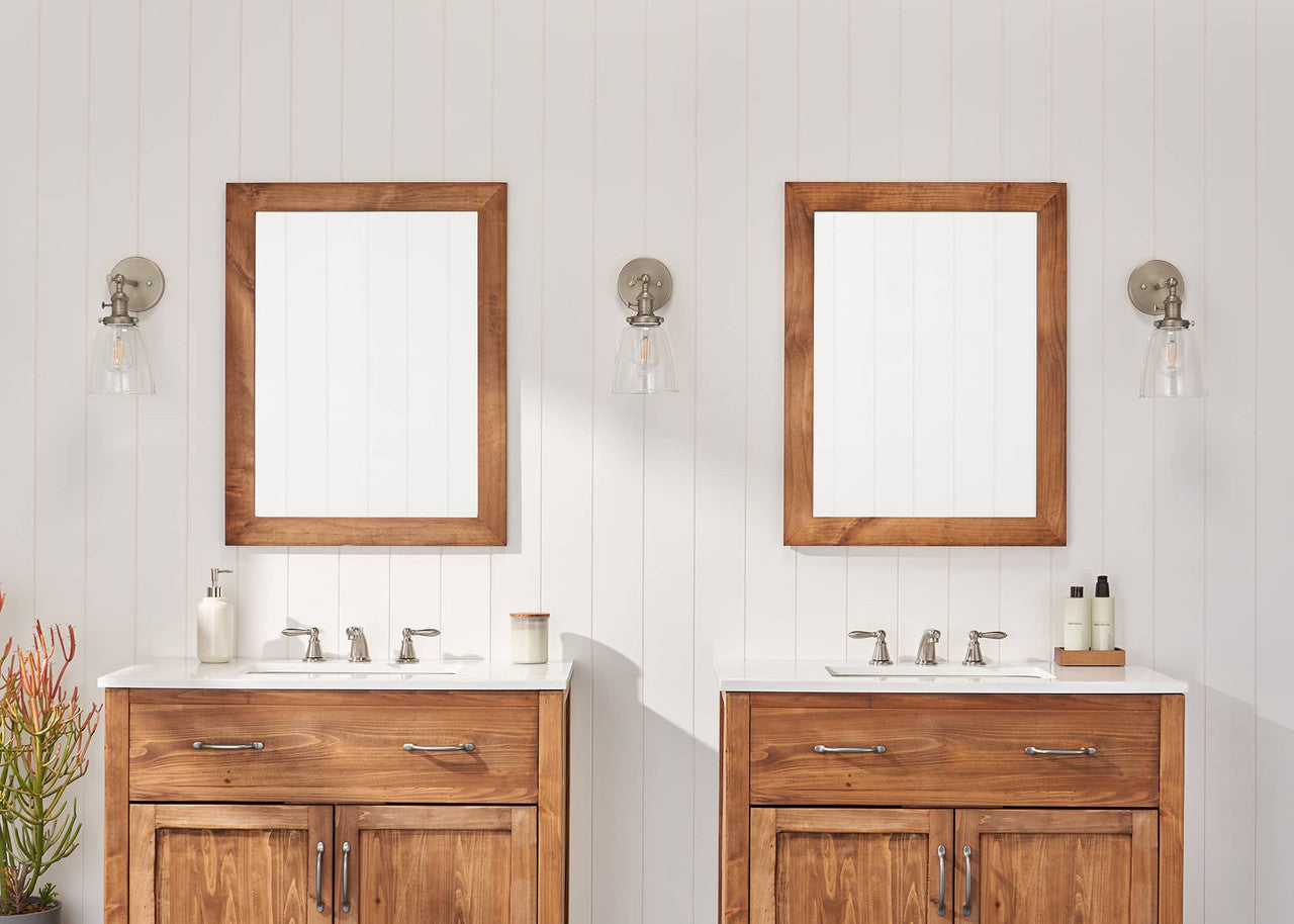 A bathroom with a double vanity setup features two wooden-framed mirrors above matching wooden cabinets with white countertops and silver faucets. Wall-mounted glass sconces provide lighting, and decorative items like soap dispensers and plants add a stylish touch.
