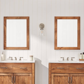 A bathroom with a double vanity setup features two wooden-framed mirrors above matching wooden cabinets with white countertops and silver faucets. Wall-mounted glass sconces provide lighting, and decorative items like soap dispensers and plants add a stylish touch.