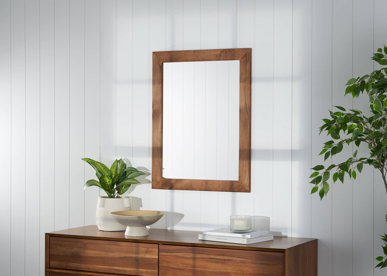 A wooden-framed rectangular mirror hangs on a white paneled wall, reflecting soft natural light. Below, a wooden dresser holds a potted plant, a ceramic bowl, a candle, and stacked books, creating a minimalist and cozy decor.
