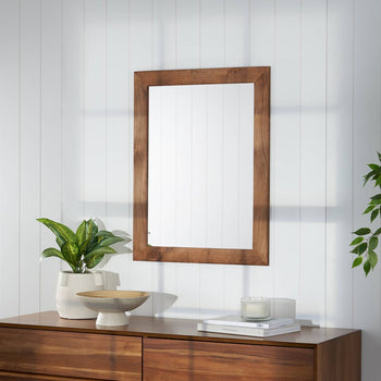 A wooden-framed rectangular mirror hangs on a white paneled wall, reflecting soft natural light. Below, a wooden dresser holds a potted plant, a ceramic bowl, a candle, and stacked books, creating a minimalist and cozy decor.