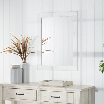 A vertical rectangle mirror with a white patterned frame is mounted on a white plank wall. Below, a light wood console table with two drawers is decorated with tall vases and a small tray.