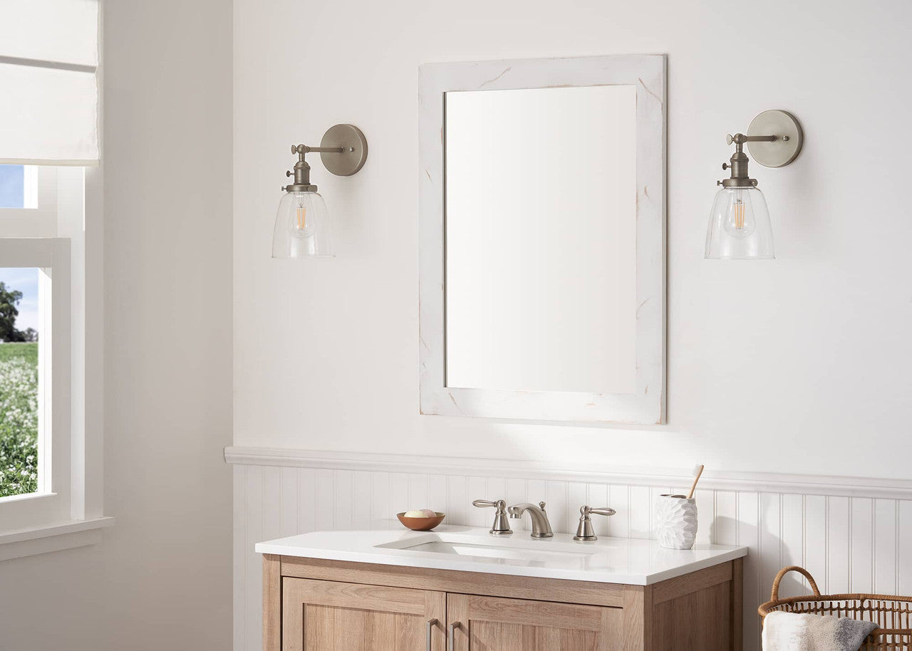 A rectangular bathroom mirror with a white marble frame hangs above a wooden vanity with a white countertop. Two wall sconces flank the mirror, and a window is visible on the left.