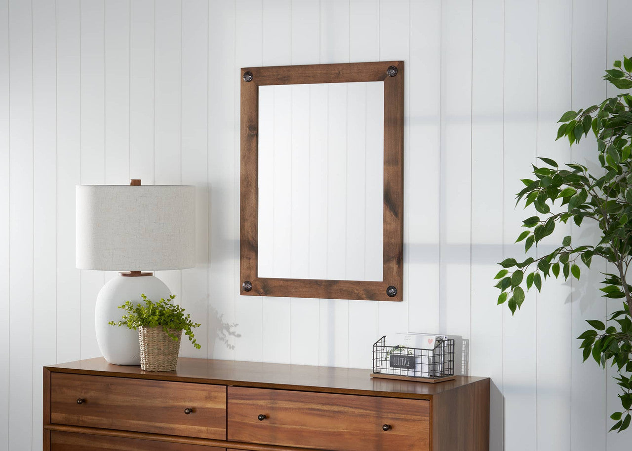 This is an eye-level, straight-on shot of a rectangular farmhouse vanity mirror with clavos in walnut, hung on a white wall with vertical paneling. The mirror is centered in the frame, with a portion of a brown wood dresser, a white lamp with a beige shade, and a plant in a woven basket visible at the bottom, and a portion of a green artificial tree on the right.