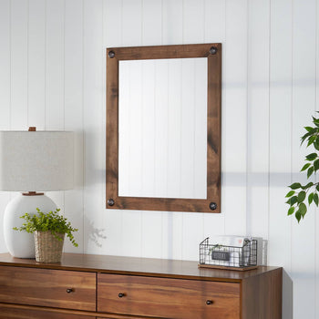 This is an eye-level, straight-on shot of a rectangular farmhouse vanity mirror with clavos in walnut, hung on a white wall with vertical paneling. The mirror is centered in the frame, with a portion of a brown wood dresser, a white lamp with a beige shade, and a plant in a woven basket visible at the bottom, and a portion of a green artificial tree on the right.