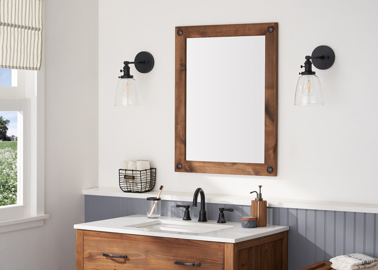 This is an eye-level, straight-on shot of a bathroom vanity area, showcasing a dark wood framed mirror with clavos (decorative nails) as the central focal point. The mirror reflects a bright window, and is flanked by two black sconces with clear glass shades, adding a blend of rustic and modern elements to the space.