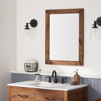This is an eye-level, straight-on shot of a bathroom vanity area, showcasing a dark wood framed mirror with clavos (decorative nails) as the central focal point. The mirror reflects a bright window, and is flanked by two black sconces with clear glass shades, adding a blend of rustic and modern elements to the space.