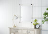 A rectangular white-framed mirror with dark round accents at each corner hangs on a white paneled wall above a light-colored wooden console table. On the table are a brass-toned lamp with a white shade, a small potted plant with white flowers, and a round black-framed clock with white numbers.