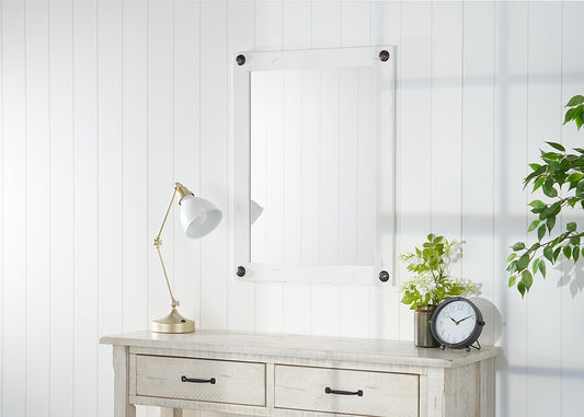 A rectangular white-framed mirror with dark round accents at each corner hangs on a white paneled wall above a light-colored wooden console table. On the table are a brass-toned lamp with a white shade, a small potted plant with white flowers, and a round black-framed clock with white numbers.
