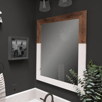 A square, two-toned farmhouse vanity mirror with a wood frame top and white painted bottom hangs on a dark gray wall above a white marble sink. The mirror reflects a slightly obscured view of the bathroom.